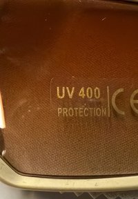 Nahaufnahme eines Objektivs mit goldenem Text „UV 400 Schutz“ und CE-Kennzeichen auf braunem, strukturiertem Hintergrund.