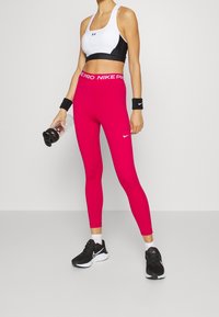 Czarny i biały stanik sportowy, różowe legginsy z logo Nike oraz czarne buty sportowe. Akcesoria to opaska na nadgarstek i butelka wody.