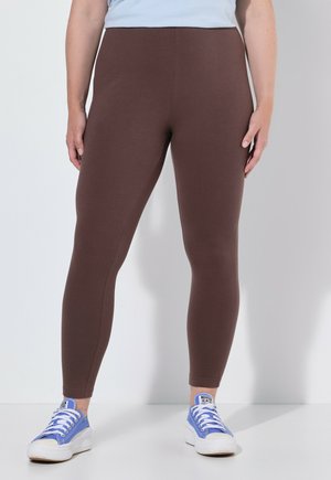 Person trägt braune enganliegende Leggings und hellblaue Sneakers und steht vor einem einfarbigen hellen Hintergrund.