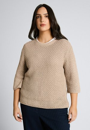 Vrouw met lang donker haar, gekleed in een beige gebreide trui met een textuur en ellebooglange mouwen, en donker gestreepte broek met krijtstrepen.