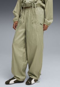 Pantaloni larghi di un verde oliva chiaro con elastico in vita, cordoncino e tasche laterali a fessura. Presenta un logo Puma nero sulla coscia.