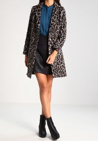 Cappotto con stampa leopardata, colletto, maniche lunghe e taglio dritto. Indossato sopra un top teal e una mini gonna nera, abbinato a stivali neri alla caviglia.