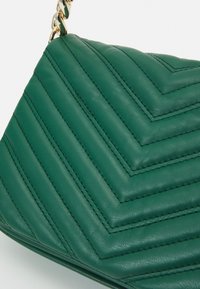 Zielona skórzana torebka z wzorem w zygzak, przeszyciami i złotym łańcuszkiem. Gładka faktura oraz wyraźnie zdefiniowany kształt.