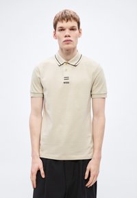 Μπεζ πόλο μπλούζα με μ�αύρο περίγραμμα και κλείσιμο με τρία κουμπιά· διαθέτει λογότυπο 'BOSS' σε μαύρο, κοντά μανίκια και υφή υφάσματος.