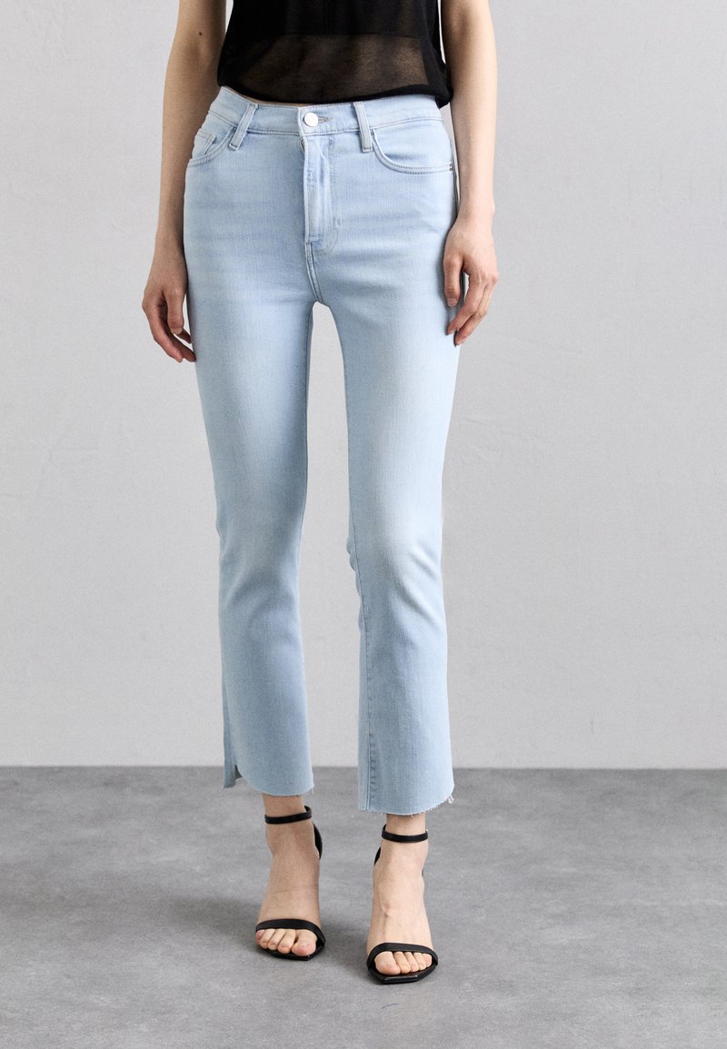 Lys blå høy midje cropped jeans med frayed kant og slisser på siden, kombinert med svarte stroppehæler på en grå bakgrunn.