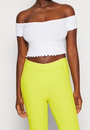 Kvinde iført hvid off-shoulder cropped top med flæsekant og lyse gule, teksturerede højtaljede bukser mod en ensfarvet baggrund.