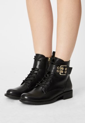 Bottes noires en cuir à la cheville avec laçage à l'avant, fermeture éclair sur le côté et large sangle dotée d'une boucle dorée portées sur des jambes nues.