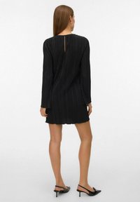 Czarna plisowana sukienka z długimi rękawami, z tylnym detalem w formie klucza i falbaniastym wykończeniem; zestawiona z czółenkami na obcasie z paskiem na pięcie.