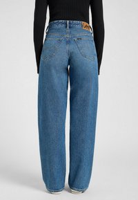 Weite blaue Denim-Jeans mit hoher Taille, mit einem braunen Lederlogo-Patch auf der Rückseite und dezenten Nähten an den Taschen.