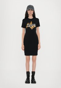 Crna majica haljina s zlatnim grafičkim printom i natpisom "VERSACE JEANS COUTURE"; uparena s crnim čizmama na vezivanje i sivom kapom.