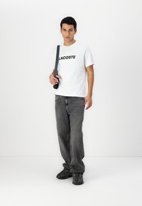 Balta medvilninė marškinėliai su juodu "LACOSTE" logotipu, derinami su laisvais pilkais džinsais ir juodais raštuotais bateliais. Modelis laiko juodą rankinę per petį.