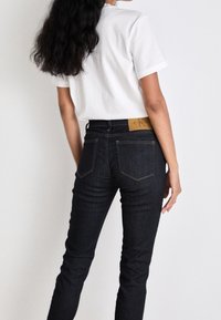 Mörka denimjeans med slim fit, har guldstickning, bakfickor och en ljusbrun läderlapp vid midjan. Vit t-shirt bärs.