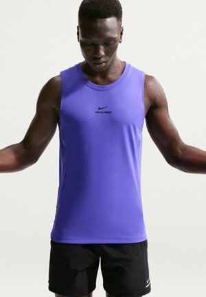 Umęczona osoba w fioletowym bezrękawniku Nike Pro i czarnych szortach Nike, patrząca w dół z lekko wyciągniętymi ramionami.