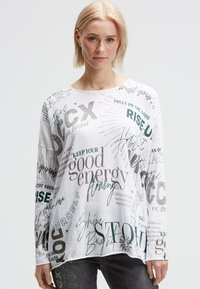 Frau mit kurzen blonden Haaren trägt ein weißes Langarmshirt mit verschiedenen motivierenden Sprüchen in unterschiedlichen Schriftarten und dunklen Jeans.