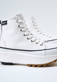 Białe trampki typu high-top na grubej białej platformie, z czarnymi akcentami i srebrnymi oczkami. Na boku znajduje się metka z logo.