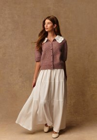 Gebreide cardigan in zacht mauve met een witte kanten kraag, knoopsluiting aan de voorkant, gecombineerd met een vloeiende witte maxi rok met lagen. Gladde textuur, ontspannen pasvorm.