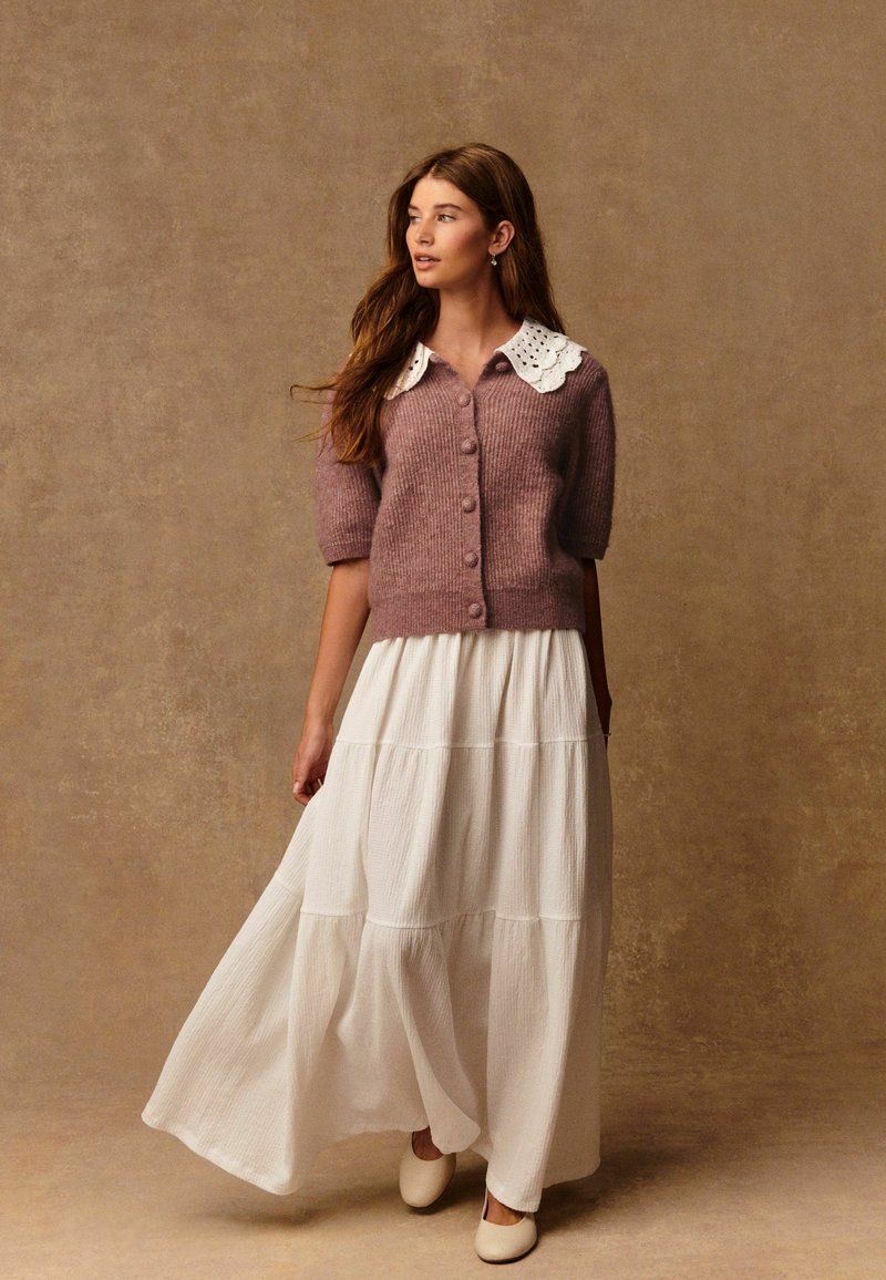 Gebreide cardigan in zacht mauve met een witte kanten kraag, knoopsluiting aan de voorkant, gecombineerd met een vloeiende witte maxi rok met lagen. Gladde textuur, ontspannen pasvorm.