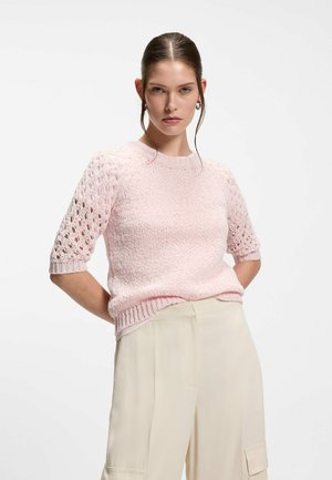 Woman with hair tied back wearing light pink textured knit short sleeve sweater and beige high-waist trousers, standing with hands behind back.