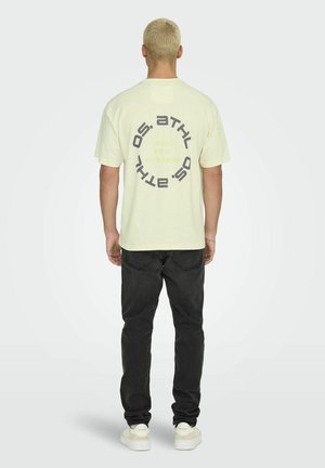 Light yellow cotton t-shirt with circular printed text on the back. Paired with black jeans and white shoes. Standard fit, short sleeves.