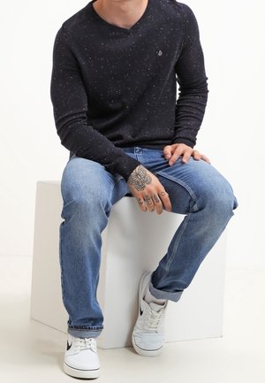 Personne assise portant un t-shirt à manches longues noir tacheté, un jean bleu clair, des baskets blanches Nike, avec des tatouages visibles sur les mains et les doigts.