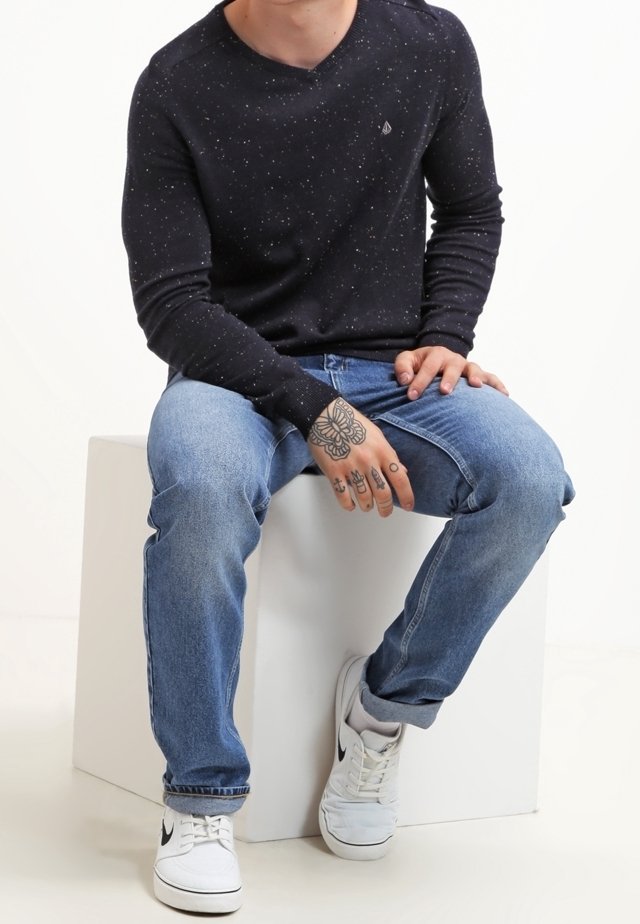 Personne assise portant un t-shirt à manches longues noir tacheté, un jean bleu clair, des baskets blanches Nike, avec des tatouages visibles sur les mains et les doigts.