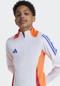 Weißer sportlicher Pullover mit orangefarbenen Akzenten, blauen Streifen auf den Schultern und einem halben Reißverschlusskragen; aus atmungsaktivem Material.