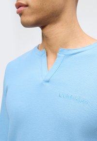 Κλείσιμο πλάνου ενός ατόμου που φοράει γαλάζιο υφασμάτινο πουκάμισο Calvin Klein με λαιμόκοψη σε σχήμα εγκοπής, εμφανίζοντας τον λαιμό και το κάτω μέρος του προσώπου.