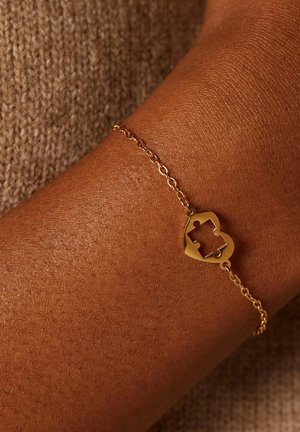 Gouden kettingarmband met een hartvormige puzzelstuk charme, met een gladde afwerking, bevestigd aan een pols tegen een achtergrond van gestructureerde bruine stof.