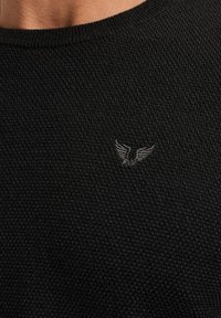 Pull en tricot noir avec un motif texturé, col rond et un petit logo d'aigle brodé en gris sur la poitrine.