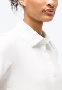 Γυναίκα με σγουρά μαλλιά φοράει άσπρο πουκάμισο με γιακά και μερικώς ορατή σειρά κουμπιών σε ανοιχτό φόντο.