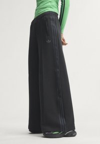 Person wearing black wide-leg Adidas pants with three black stripes, green long-sleeve top, and green-black sneakers standing sideways.