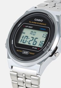 Montre Casio numérique avec un cadran noir, un bracelet en acier inoxydable argenté et un affichage LCD clair montrant l'heure, la date et les réglages d'alarme. Imperméable.