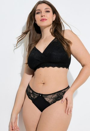 Femme portant un bralette en dentelle noire et une culotte assortie bordée de dentelle, debout devant un fond blanc.