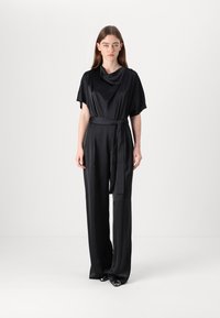 Svart sateng jumpsuit med vide ermer, en bølget hals, en figursydd midje med belte, og flytende, rette benbukser.