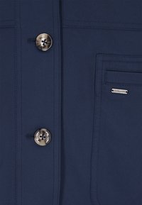 Tengerészkék zakó két fényes ezüstszínű gombbal, mellkasi zsebbel és finom mintázattal, varrási részletekkel.