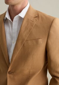Blazer in lino marrone con revers, chiusura a un bottone e taschino sul petto, abbinato a una camicia in lino bianca sottostante.