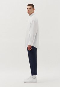 Wit gestreepte longsleeve shirt met een zak, gecombineerd met een marineblauwe broek en witte sneakers. Model staat in profiel tegen een effen achtergrond.