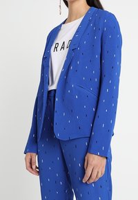 Modrý blejzr s bílým a černým tečkovaným vzorem, přiléhavý střih a zapínání na jeden knoflík. V kombinaci s odpovídajícími kalhotami. Hladká textura látky.