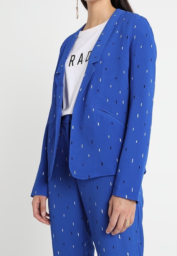 Modrý blejzr s bílým a černým tečkovaným vzorem, přiléhavý střih a zapínání na jeden knoflík. V kombinaci s odpovídajícími kalhotami. Hladká textura látky.