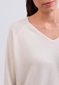 Pullover in maglina color beige chiaro con scollatura a V. Presenta maniche raglan e una texture morbida, evidenziando una vestibilità rilassata e dettagli di design minimali.