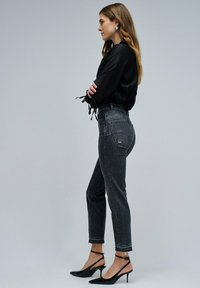 Blusa negra con mangas atadas, jeans de cintura alta en gris oscuro y tacones negros de punta. Diseño simple, silueta ajustada y puños deshilachados.