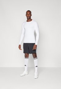 Weißes langärmliges Sportoberteil, schwarze Shorts mit seitlichen Streifen, weiße Socken mit schwarzen Streifen und Logo sowie weiße Fußballschuhe.