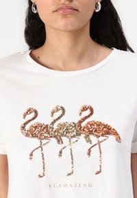Hvid t-shirt med tre flamingoer lavet af flerfarvede paljetter, med en tekst nedenunder der læser "FLAMAZING", i et afslappet design.