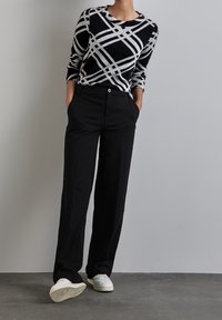 Osoba ubrana w czarne spodnie, sweter w geometryczny czarno-biały wzór, białe trampki, stojąca z rękami w kieszeniach na tle szarej ściany.