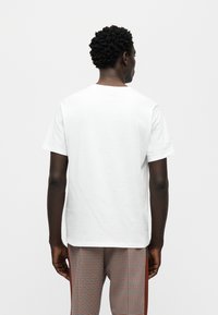 T-shirt blanc en coton à col rond, vu de dos. Porté avec un pantalon à motif à carreaux avec des bandes latérales rouges.