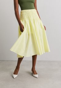 Jupe jaune clair, longueur genoux, avec une taille smockée et un tissu fluide. Portée avec un haut vert et des escarpins blancs à bout pointu.