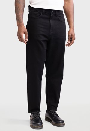 Mężczyzna noszący czarne jeansy o prostych nogawkach, czarne sznurowane buty z żółtym przeszyciem oraz biały top bez rękawów, stojący na jasnym tle.