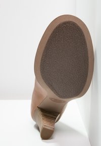 Chaussure à talon marron avec une semelle extérieure en caoutchouc texturé, dotée d'un talon en bois rayé et d'un dessus en cuir lisse, inclinée contre un fond blanc.