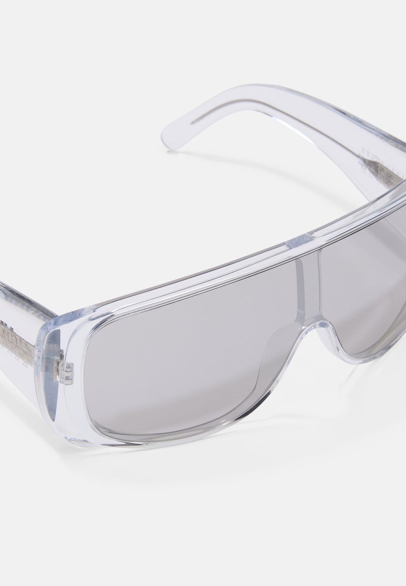 Occhiali da sole trasparenti con un design elegante e avvolgente. Presentano lenti grigie sfumate e una montatura in plastica trasparente con bordi lisci.