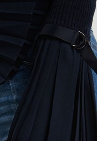 Gros plan sur un tissu plissé bleu marine avec une manche tricotée noire et une sangle ajustable avec une boucle argentée sur un jean en denim.